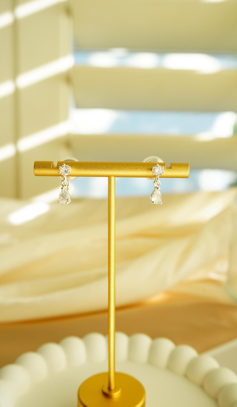 K&ouml;nnte beinhalten: Ein Paar goldfarbene Ohrringe auf einem goldenen Pr&auml;sentationsst&auml;nder. Jeder Ohrring hat einen kleinen, runden, klaren Stein oben und einen tropfenf&ouml;rmigen, klaren Stein, der darunter h&auml;ngt. Der Hintergrund umfasst einen wei&szlig;en, geriffelten Teller und weichen, verschwommenen Stoff.