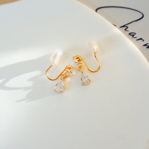 Op de afbeelding: Een paar gouden clip-oorbellen met een traanvormige kristallen hanger. De oorbellen hebben een kleine, transparante plastic clip aan de achterkant.