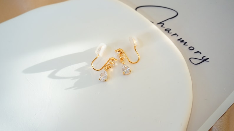 Op de afbeelding: Een paar gouden clip-oorbellen met een traanvormige kristallen hanger. De oorbellen hebben een kleine, transparante plastic clip aan de achterkant.
