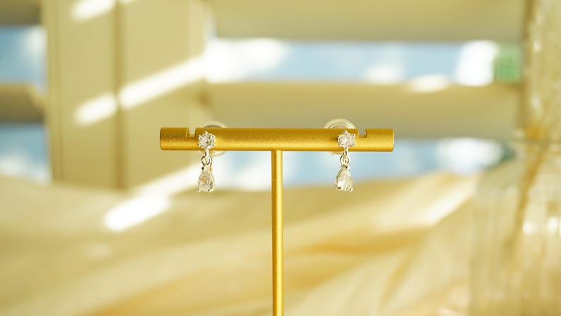 K&ouml;nnte beinhalten: Ein Paar silberfarbene Ohrringe mit einem kleinen, runden, klaren Edelstein oben und einem tropfenf&ouml;rmigen Edelstein, der darunter h&auml;ngt. Die Ohrringe werden auf einem goldfarbenen St&auml;nder vor einem weichen, verschwommenen Hintergrund pr&auml;sentiert.