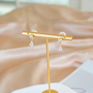 Op de afbeelding: Een paar gouden oorhangers met heldere zirkonia-stenen. De oorbellen worden gepresenteerd op een gouden standaard.