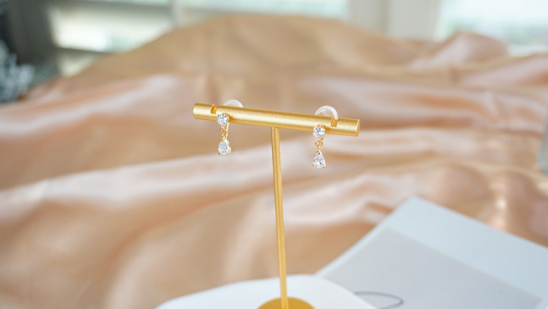 Op de afbeelding: Een paar gouden oorhangers met heldere zirkonia-stenen. De oorbellen worden gepresenteerd op een gouden standaard.