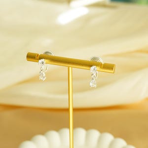 K&ouml;nnte beinhalten: Ein Paar silberfarbene H&auml;ngeohrringe mit klaren, tropfenf&ouml;rmigen Steinen werden auf einem goldfarbenen Schmuckst&auml;nder pr&auml;sentiert. Die Ohrringe sind an einem kleinen, runden Stecker befestigt. Der St&auml;nder steht auf einer wei&szlig;en, gewellten Schale.