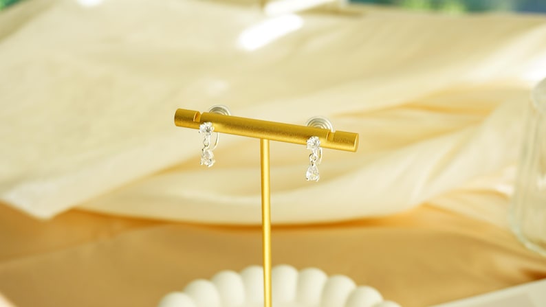 K&ouml;nnte beinhalten: Ein Paar silberfarbene H&auml;ngeohrringe mit klaren, tropfenf&ouml;rmigen Steinen werden auf einem goldfarbenen Schmuckst&auml;nder pr&auml;sentiert. Die Ohrringe sind an einem kleinen, runden Stecker befestigt. Der St&auml;nder steht auf einer wei&szlig;en, gewellten Schale.