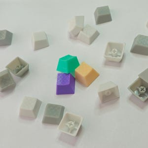 Può includere: Una collezione di tasti colorati per tastiera sparsi su una superficie bianca. I tasti sono di vari colori, tra cui verde, viola e beige, alcuni con superfici strutturate. Alcuni tasti sono staccati, rivelando i loro meccanismi interni.