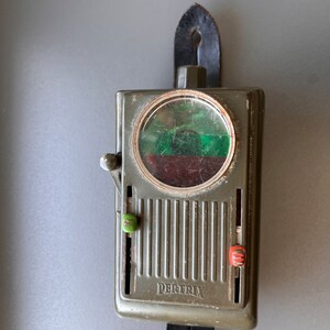 May include: A green metal device with a round window and two knobs. The device is labeled "DERTRIX" and has a black leather strap attached.