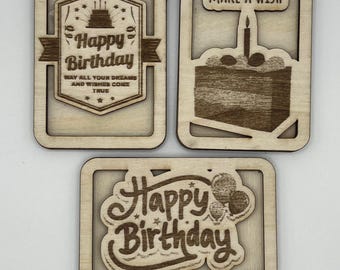 May include: Three wooden birthday cards with laser-cut designs. The first card says "Happy Birthday" with a cake and stars. The second card says "Make a Wish" with a cake and a candle. The third card says "Happy Birthday" with balloons.