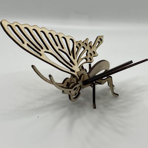 Puede incluir: Un modelo de mariposa de madera con detalles intrincados, elaborado en madera marrón claro. Las alas están extendidas y el cuerpo está detallado con patas y antenas.