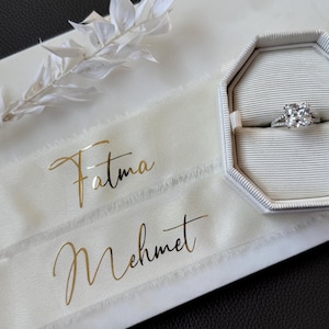 Könnte beinhalten: Zwei weiße Satinbänder mit goldener Beschriftung. Das obere Band sagt "Fatma" und das untere Band sagt "Mehmet". Im Hintergrund befindet sich eine weiße Ringbox mit einem silbernen Ring mit einem großen, klaren Stein.