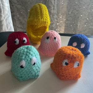 May include: A set of seven crocheted Pac-Man ghosts in various colors, including red, pink, blue, green, orange, and yellow. The ghosts are all in a classic Pac-Man style, with round bodies and large eyes.