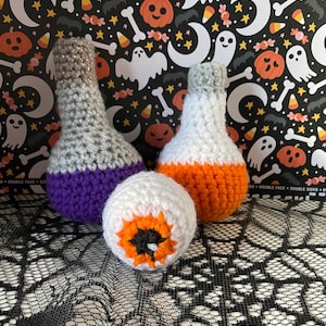 May include: Two crocheted potion bottles, one purple and one orange, with a crocheted eyeball in the center. The bottles and eyeball are on a black lace background.