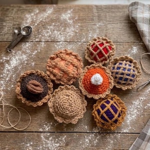 Puede incluir: Una colección de siete tartas en miniatura tejidas a ganchillo en varios colores, incluyendo rojo, naranja, azul, morado, marrón y marrón oscuro, con costras de rejilla y tapas sólidas, dispuestas sobre una superficie de madera con cucharas medidoras.