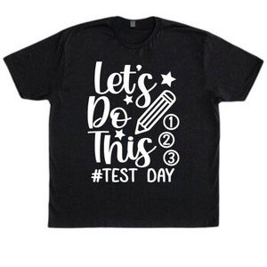 May include: Black t-shirt with white text that reads "Let's Do This 1 2 3 #Test Day" with a pencil and stars.