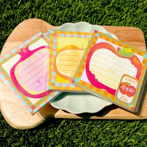 May include: Three colorful fruit-themed sticky note pads on a wooden cutting board. The pads feature apple, lemon, and pomegranate designs with lined spaces for notes. The apple pad has a label that reads "apple" and "8.5 cm 50 sheets".