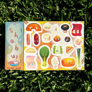 Puede incluir: Una hoja de pegatinas y una tarjeta con ilustraciones coloridas de alimentos. La hoja de pegatinas incluye varias verduras y otros alimentos con caras bonitas. La tarjeta dice "shabui" y "sticker sheet".