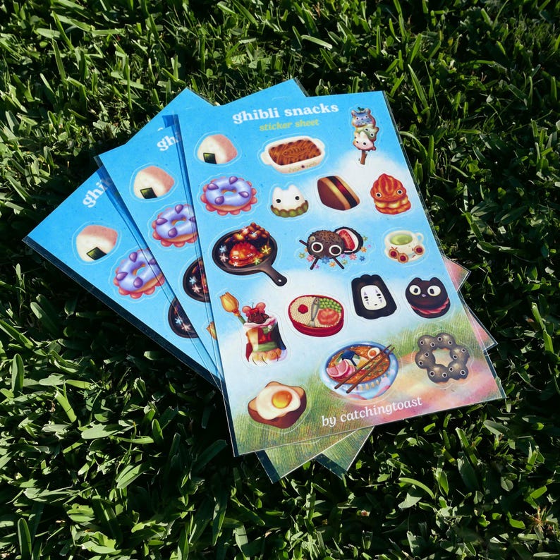 Puede incluir: Hojas de pegatinas con ilustraciones coloridas de varios bocadillos y personajes, con el texto "ghibli snacks sticker sheet" y "by catchingtoast" sobre un fondo azul. Las pegatinas est&aacute;n dispuestas sobre un lecho de hierba verde.