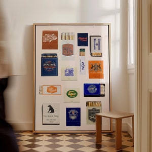 May include: A framed collage of matchbox labels featuring various designs and text, including "The Black Dog", "River City", "Heritage Classic", and "Edmonton Oilers". The collage is displayed on a white wall with a wooden floor.