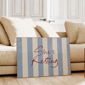 May include: A blue and white striped poster with the text "She's Resting" in red cursive font. The poster is leaning against a beige sofa with cushions.