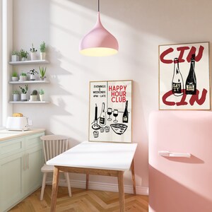 Op de afbeelding: Een roze retro koelkast met een witte handgreep, naast een witte tafel met een houten onderstel. Twee ingelijste posters hangen aan de muur achter de koelkast. Een poster zegt "Happy Hour Club" en de andere zegt "Cin".