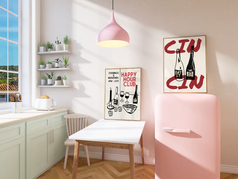 Op de afbeelding: Een roze retro koelkast met een witte handgreep, naast een witte tafel met een houten onderstel. Twee ingelijste posters hangen aan de muur achter de koelkast. Een poster zegt "Happy Hour Club" en de andere zegt "Cin".