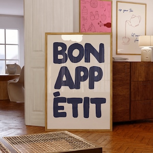 May include: A white framed poster with the text "BON APPÉTIT" in navy blue lettering. The poster is hanging on a white wall in a room with wooden floors.