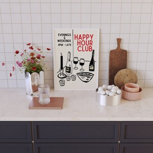 Op de afbeelding: Een wit ingelijst print met zwarte tekst die "Happy Hour Club" en "Evenings & Weekends 4PM - Late" luidt. De print toont een zwart-witte illustratie van wijnglazen, flessen en een kom met snacks. De print staat op een wit aanrecht met een vaas met roze bloemen, een glas, een boek, een kom met witte stenen en een roze kom.