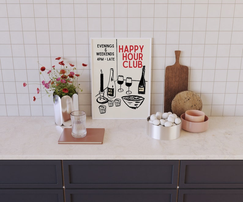 Op de afbeelding: Een wit ingelijst print met zwarte tekst die "Happy Hour Club" en "Evenings & Weekends 4PM - Late" luidt. De print toont een zwart-witte illustratie van wijnglazen, flessen en een kom met snacks. De print staat op een wit aanrecht met een vaas met roze bloemen, een glas, een boek, een kom met witte stenen en een roze kom.