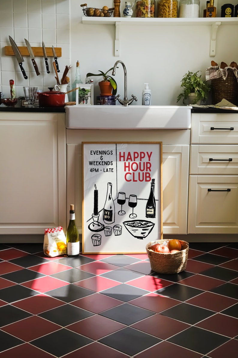 Op de afbeelding: Een ingelijst print met de tekst "Happy Hour Club" in rode en zwarte letters. De print toont een zwart-witte illustratie van wijnglazen, een fles wijn en een kom met eten. De print hangt aan een witte muur in een keuken.
