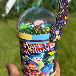 May include: A cylindrical container with a clear dome lid, filled with colorful trinkets and a character figurine. The container is decorated with the words "Happy Birthday" and character illustrations. Attached are three circular decorations with colorful polka dots and a character image.