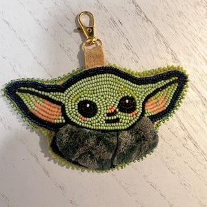 Pu&ograve; includere: Un portachiavi in perline raffigurante un personaggio stilizzato con grandi occhi scuri, pelle verde chiaro e orecchie interne color pesca. Il design &egrave; bordato da perline blu scuro con una consistenza grigia soffice alla base e un moschettone dorato.