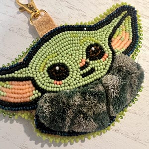 May include: A beaded keyring featuring a character with large dark eyes, light green skin, and peach-coloured inner ears. The design is outlined in dark blue beads and includes a textured, dark green fuzzy fabric accent. A gold-tone metal lobster clasp is attached to the top with a tan fabric loop.