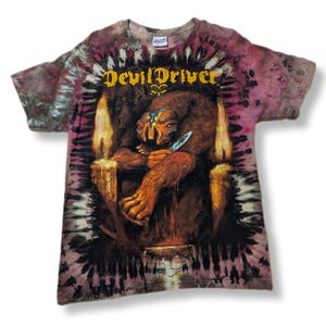 May include: Tie-dye t-shirt featuring the band name "DevilDriver" in gold lettering. The shirt displays a detailed illustration of a creature holding a knife, flanked by lit candles. The shirt's colors include shades of purple, brown, and gold.