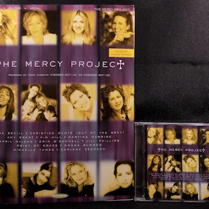 May include: The Mercy Project album cover featuring a group of women singers. The album cover is purple and gold with a cross in the top left corner. The album title is "The Mercy Project" and the album is recorded on word cassette F080668 607142, CD F080608 607128. The album features Lisa Bevill, Christine Dente, Amy Grant, Kim Hill, Martina McBride, April McLean, Erin O'Donnell, Jill Phillips, Point of Grace, Donna Summer, Michelle Tumes, and Darlene Zschech.