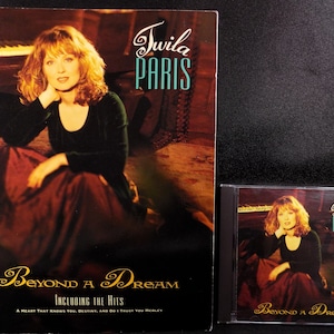 May include: A CD and a booklet for the album "Beyond a Dream" by singer Twila Paris. The cover art features a woman in a burgundy dress sitting on a rug with a piano in the background.