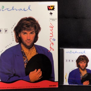 May include: A white CD case with a blue and white image of a man holding a black hat. The text "michael smith" is written in blue on the case. The text "Go West Young Man" is written in black on the case.