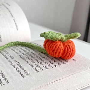 May include: A handmade crochet bookmark shaped like an orange pumpkin with green leaves, resting on an open book. The bookmark is a unique accessory for book lovers. The book has printed text. The bookmark is a unique accessory.