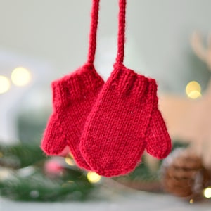 May include: A pair of red knitted mittens hanging from a string. The mittens are in focus, while the background is blurred and features a white star and a wooden reindeer.