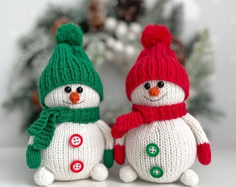 May include: Two knitted snowmen with white bodies, wearing green and red hats and scarves. Each snowman has a carrot nose, button eyes, and a stitched smile. Red and green buttons adorn their bellies. The snowmen are approximately 8 inches tall.