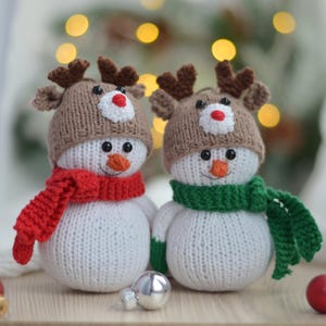 Może przedstawiać: Dwa dzianinowe ozdoby w kształcie bałwana z czapkami i szalikami renifera. Jeden bałwan ma czerwony szalik, a drugi zielony.