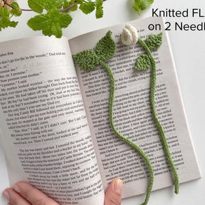 May include: A handmade knitted bookmark featuring a white flower with green leaves and a long green stem, resting on an open book page. The text Knitted FLAT on 2 Needles is printed in the upper right corner of the image.