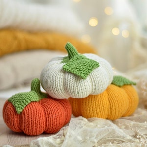 May include: Three knitted pumpkins, one white, one orange and one yellow, with green leaves. The pumpkins are sitting on a white crocheted surface.