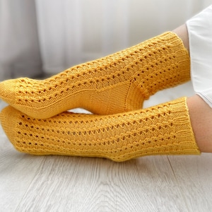 Puede incluir: Un par de calcetines de punto amarillo mostaza con un delicado calado. Los calcetines, que llegan hasta la rodilla, parecen suaves y cómodos. Se muestran sobre un suelo de madera clara.