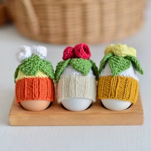 May include: Three white eggs are decorated with knitted hats. The hats are different colors and have flower designs on top. The eggs are sitting in a wooden egg carton.