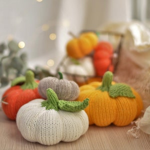 May include: Five knitted pumpkins in various colours, including orange, white, grey, and yellow. The pumpkins are arranged on a wooden surface.