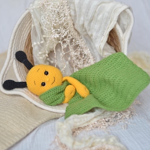 Puede incluir: Un juguete de abeja tejido a mano en amarillo y negro, con una bufanda y una manta verdes. La abeja tiene una cara sonriente y está metida en una cesta blanca con un forro color crema.