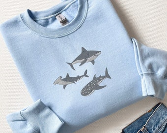May include: A light blue sweatshirt featuring an embroidered design of four different types of sharks. The sharks are in grey on the front of the sweatshirt.