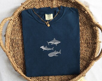 Puede incluir: Una camiseta azul marino con un diseño bordado de tiburones. La camiseta está doblada y se muestra en una cesta tejida.