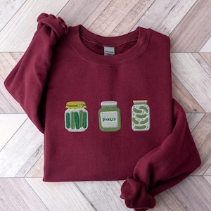 Peut inclure: Un sweat-shirt à col rond bordeaux avec une broderie représentant trois bocaux. Le bocal de gauche contient des cornichons verts verticaux, celui du centre est vert avec le texte pickles, et celui de droite contient des cornichons empilés.