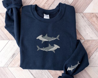 May include: A navy blue crewneck sweatshirt featuring an embroidered design of two grey hammerhead sharks on the chest and a single smaller embroidered grey hammerhead shark on the lower portion of the right sleeve, with a ribbed collar and cuffs.