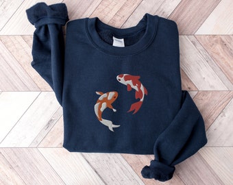 May include: A navy blue crewneck sweatshirt featuring an embroidered design of two koi fish. One fish is orange and white, while the other is red and white. The sweatshirt has long sleeves with ribbed cuffs and a ribbed neckline with a white brand label.